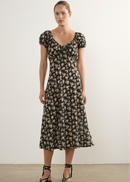 A woman stands in a studio wearing a black midi dress with a beige floral pattern, short puff sleeves, and a gathered waist. She is wearing black strappy flat sandals against a plain off-white wall and concrete floor.