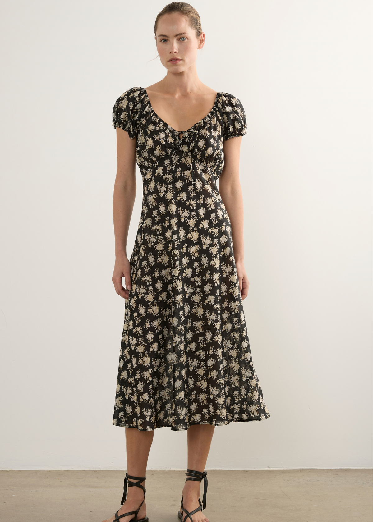 A woman stands in a studio wearing a black midi dress with a beige floral pattern, short puff sleeves, and a gathered waist. She is wearing black strappy flat sandals against a plain off-white wall and concrete floor.