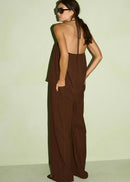 A woman wearing a dark brown ribbed halter jumpsuit with wide legs, photographed from the back against a pale yellow-green studio wall.