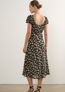 A woman modelling a black midi dress with a beige floral pattern, shown from the back. The dress features short puff sleeves, a scoop neckline, and a gathered waist.