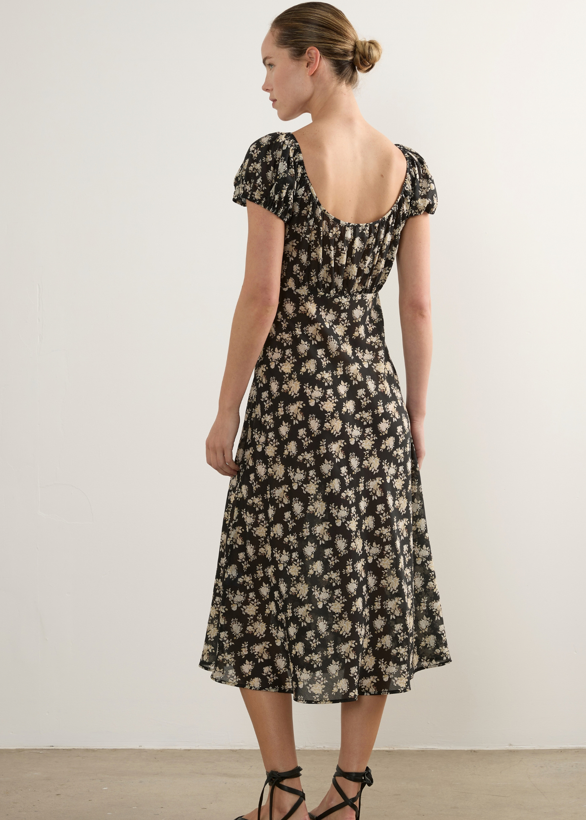 A woman modelling a black midi dress with a beige floral pattern, shown from the back. The dress features short puff sleeves, a scoop neckline, and a gathered waist.