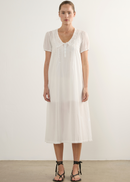 A woman modeling a white short-sleeve midi dress with lace detailing at the neckline and hem, paired with black strappy flat sandals, standing against a plain white wall.