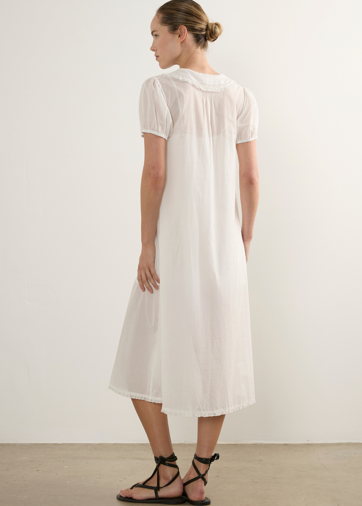 A woman wearing a short-sleeved white midi dress with a gathered neckline, shown from the back against a plain off-white wall and concrete floor.