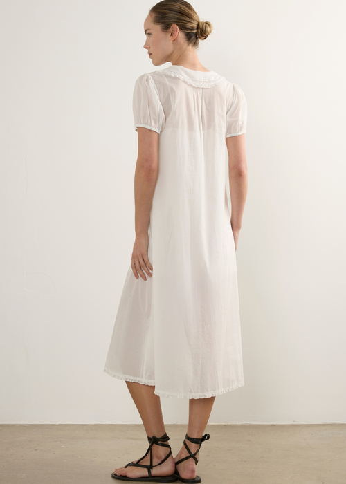 A woman wearing a short-sleeved white midi dress with a gathered neckline, shown from the back against a plain off-white wall and concrete floor.
