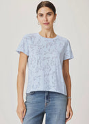 A woman viewed from the back wearing a light blue short-sleeve t-shirt with a subtle tonal leaf pattern and light-wash blue jeans, set against a plain light beige studio background.