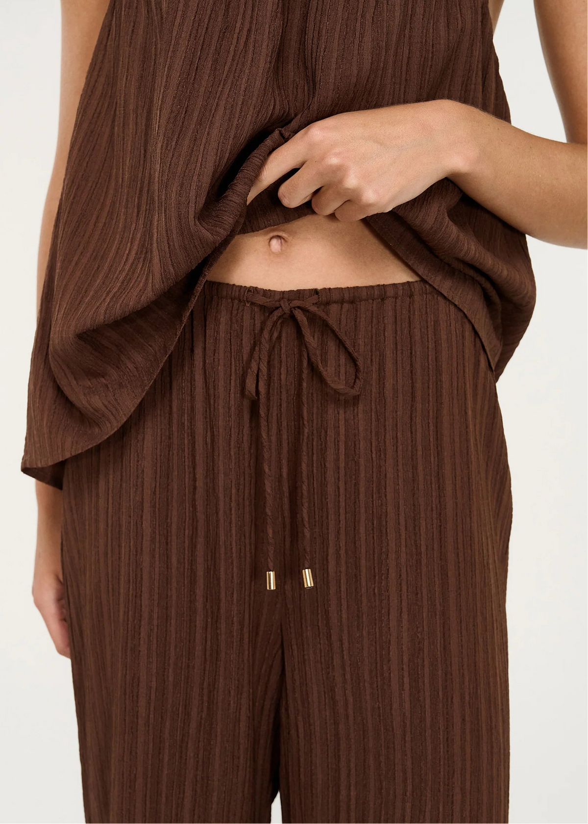 A close-up of a person wearing a brown ribbed two-piece outfit with a sleeveless top and matching drawstring pants, against a plain light background.