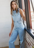 A woman wearing a light blue denim sleeveless jumpsuit with wide legs and front pockets, standing by a window in a room with white brick and wood flooring.