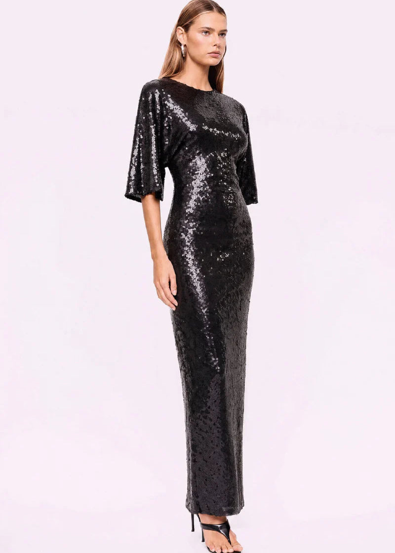 A woman modelling a floor-length black sequined gown with elbow-length flared sleeves, photographed against a plain light background.