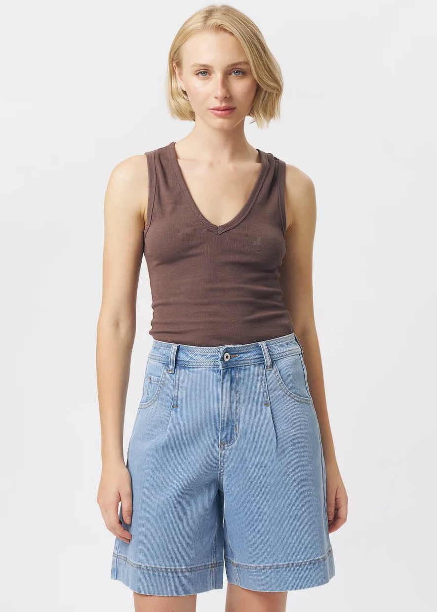 A woman modelling a brown ribbed sleeveless V-neck tank top and light-wash blue denim shorts against a plain light grey studio background.