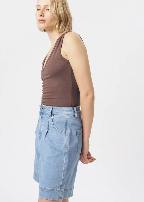 A woman modelling a brown ribbed sleeveless V-neck tank top and light-wash blue denim shorts against a plain light grey studio background.