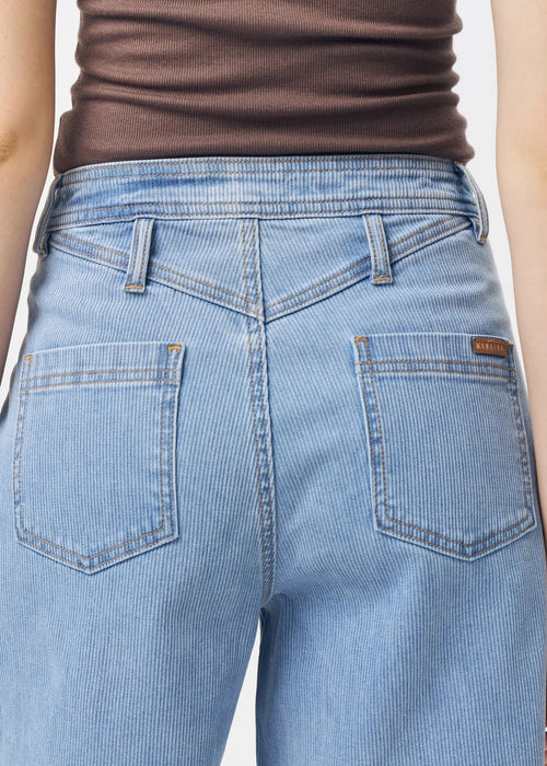 A woman modelling a brown ribbed sleeveless V-neck tank top and light-wash blue denim shorts against a plain light grey studio background.