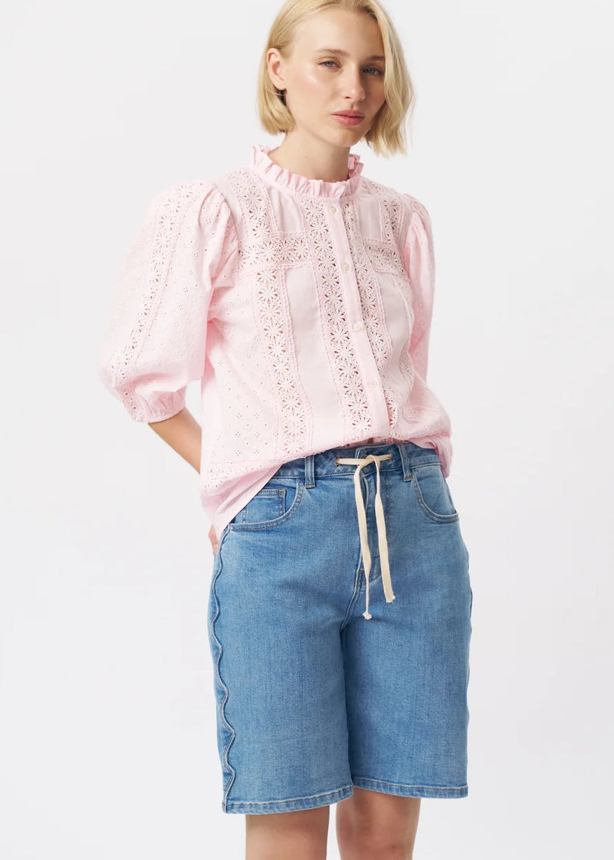 A woman modelling a light pink eyelet blouse with puff sleeves and a ruffled collar, paired with blue denim shorts, against a plain white studio background.