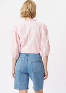 A woman modelling a light pink eyelet blouse with puff sleeves and a ruffled collar, paired with blue denim shorts, against a plain white studio background.