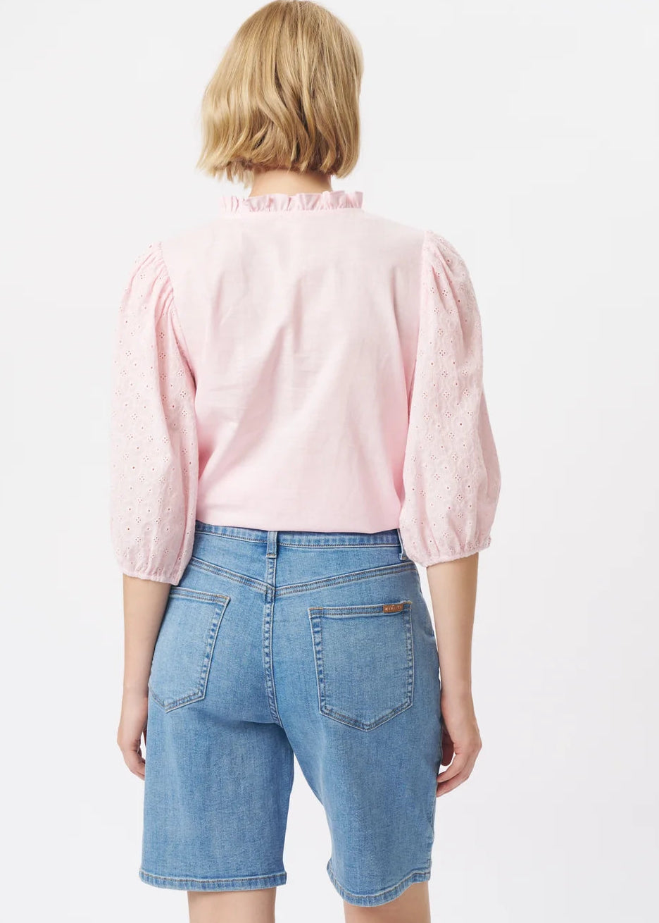 A woman modelling a light pink eyelet blouse with puff sleeves and a ruffled collar, paired with blue denim shorts, against a plain white studio background.