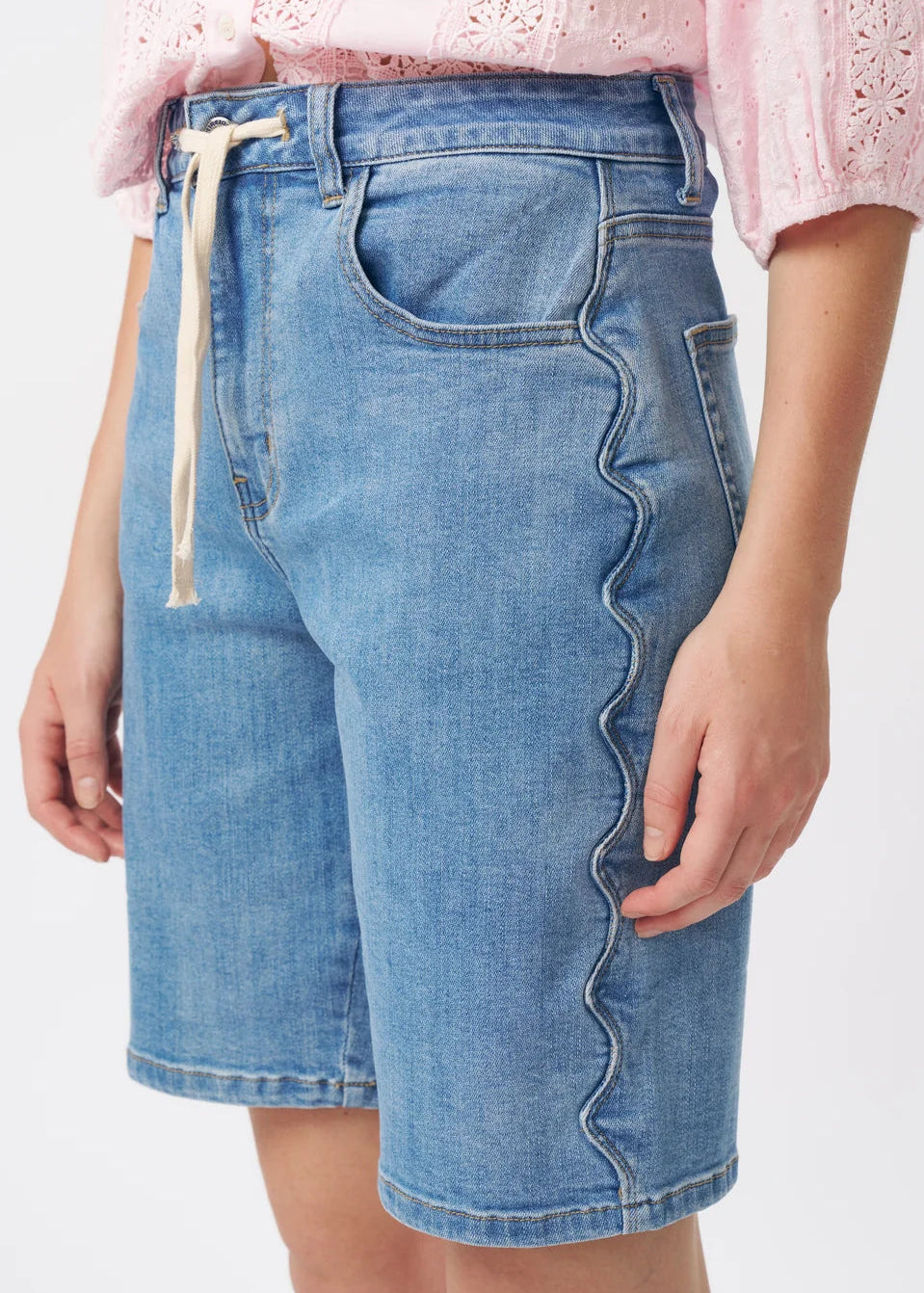 A woman modelling a light pink eyelet blouse with puff sleeves and a ruffled collar, paired with blue denim shorts, against a plain white studio background.
