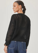 A model wearing a black open-knit cardigan with buttons and blue jeans, standing against a light grey studio background.