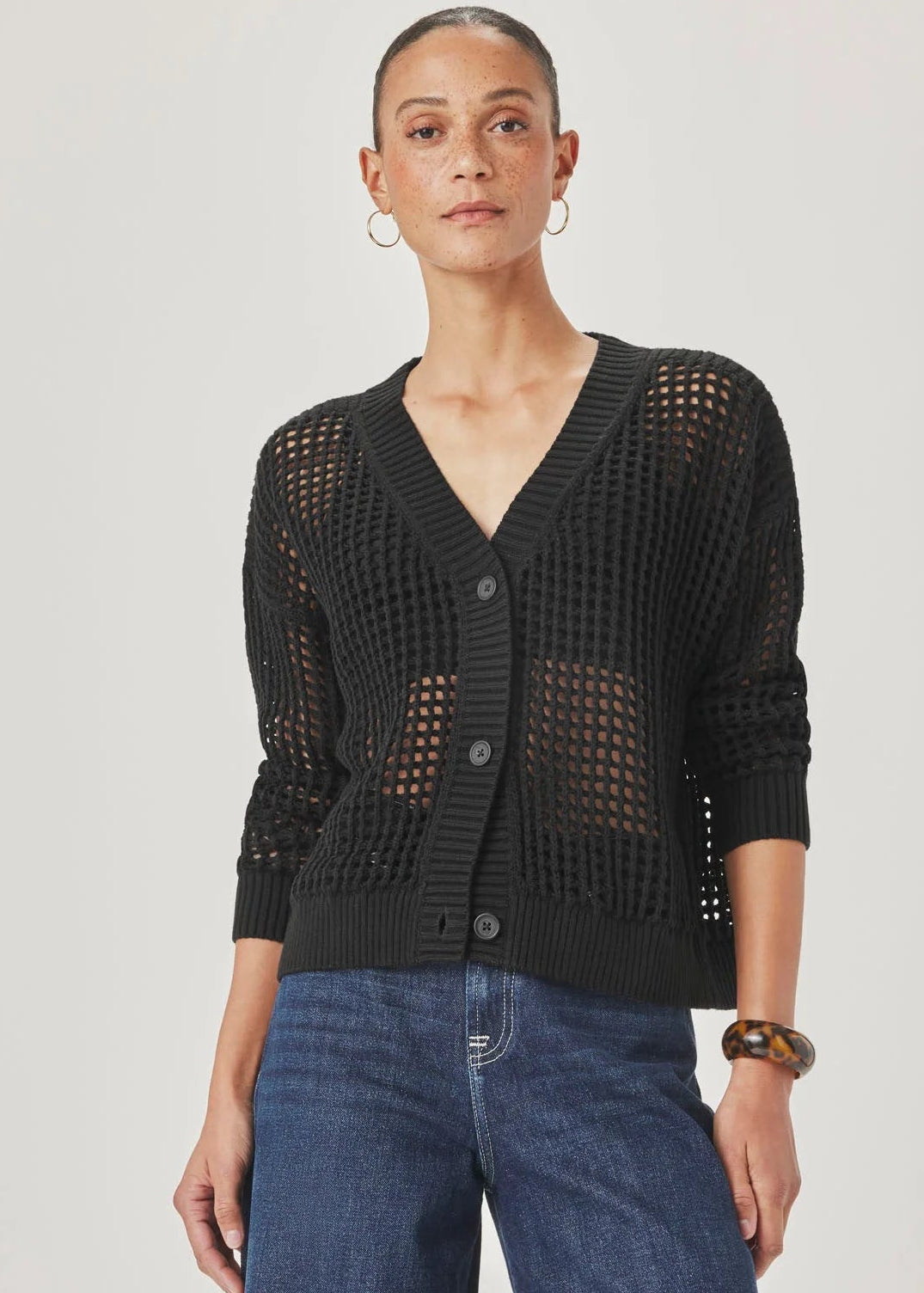 A model wearing a black open-knit cardigan with buttons and blue jeans, standing against a light grey studio background.