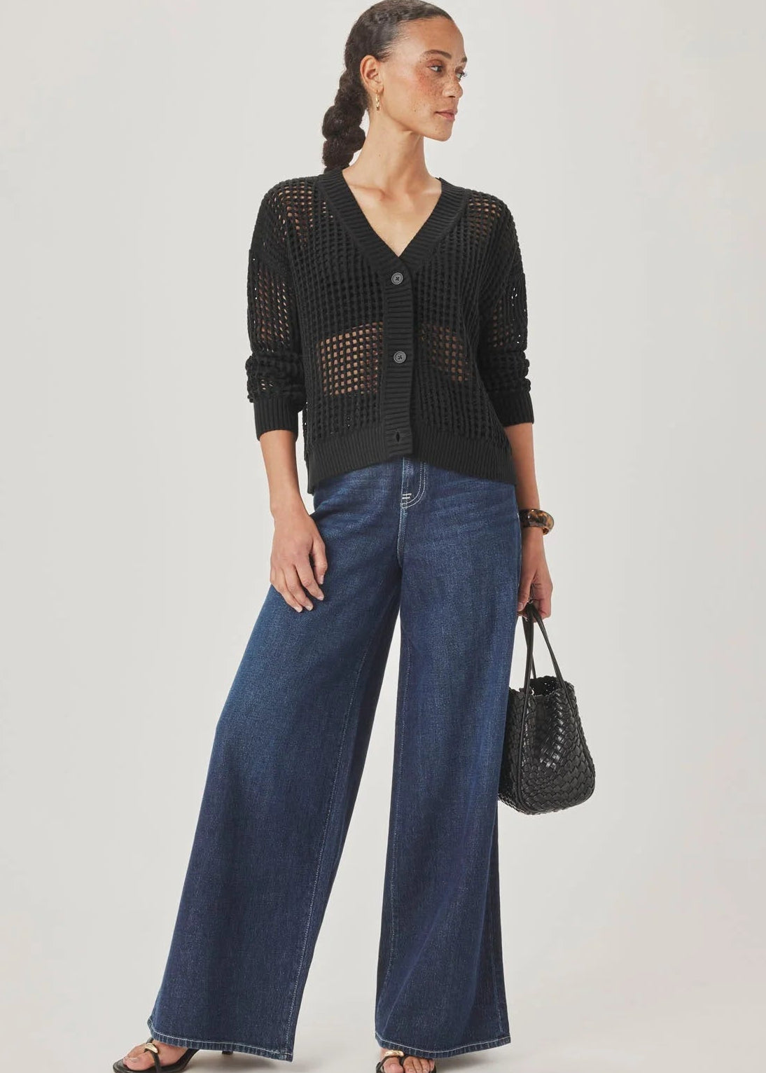 A model wearing a black open-knit cardigan with buttons and blue jeans, standing against a light grey studio background.