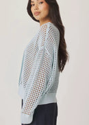 A woman modelling a light blue open-knit cardigan with a V-neck and button front, paired with dark navy pants, against a plain light background.