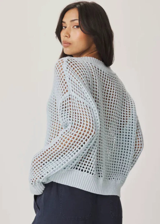 A woman modelling a light blue open-knit cardigan with a V-neck and button front, paired with dark navy pants, against a plain light background.