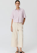 A woman modelling a light pink and white vertically striped short-sleeve button-up shirt paired with cream wide-leg cropped pants and black flip-flop sandals, set against a plain light grey studio background.