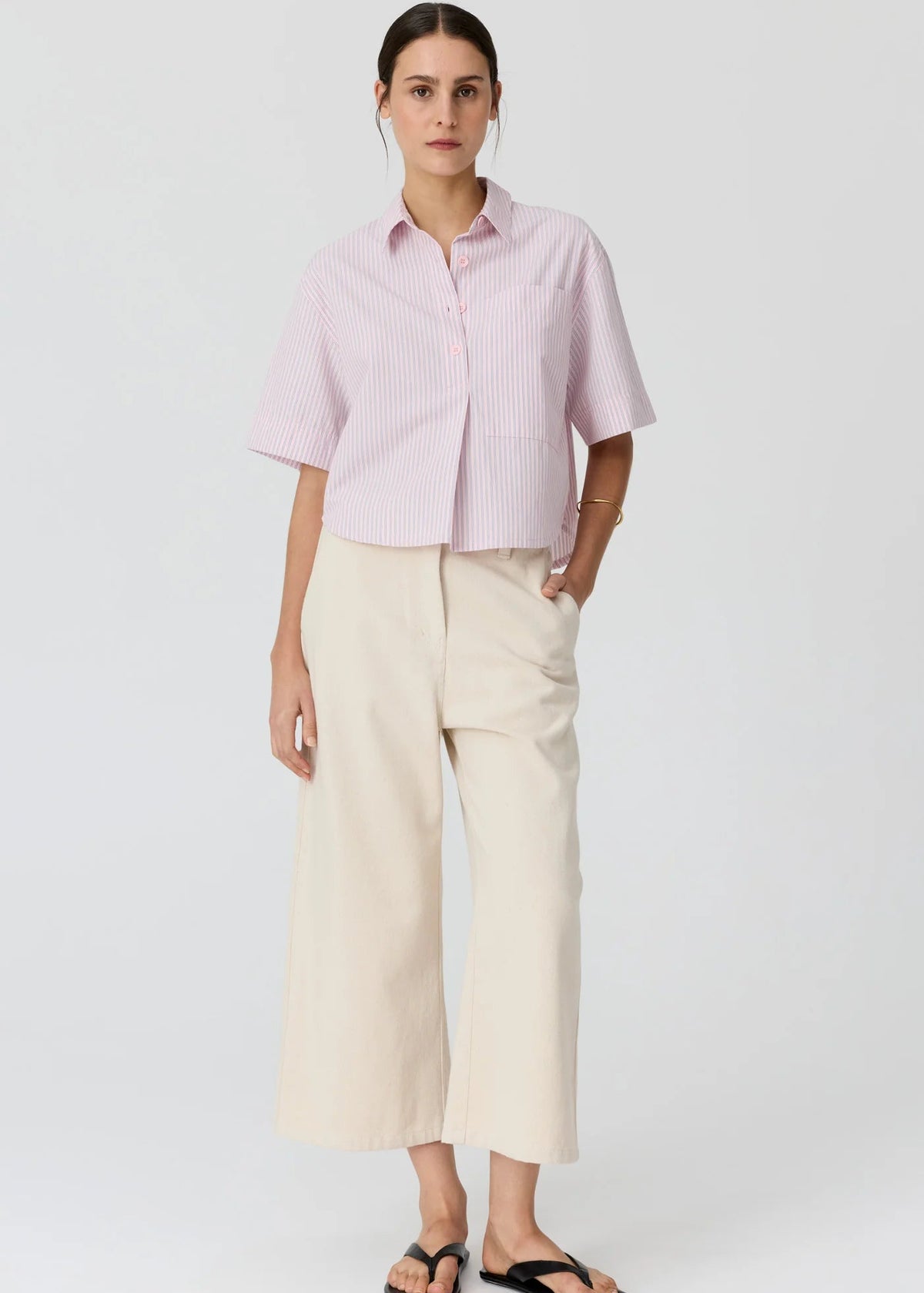 A woman modelling a light pink and white vertically striped short-sleeve button-up shirt paired with cream wide-leg cropped pants and black flip-flop sandals, set against a plain light grey studio background.