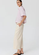 A woman modelling a light pink and white vertically striped short-sleeve button-up shirt paired with cream wide-leg cropped pants and black flip-flop sandals, set against a plain light grey studio background.