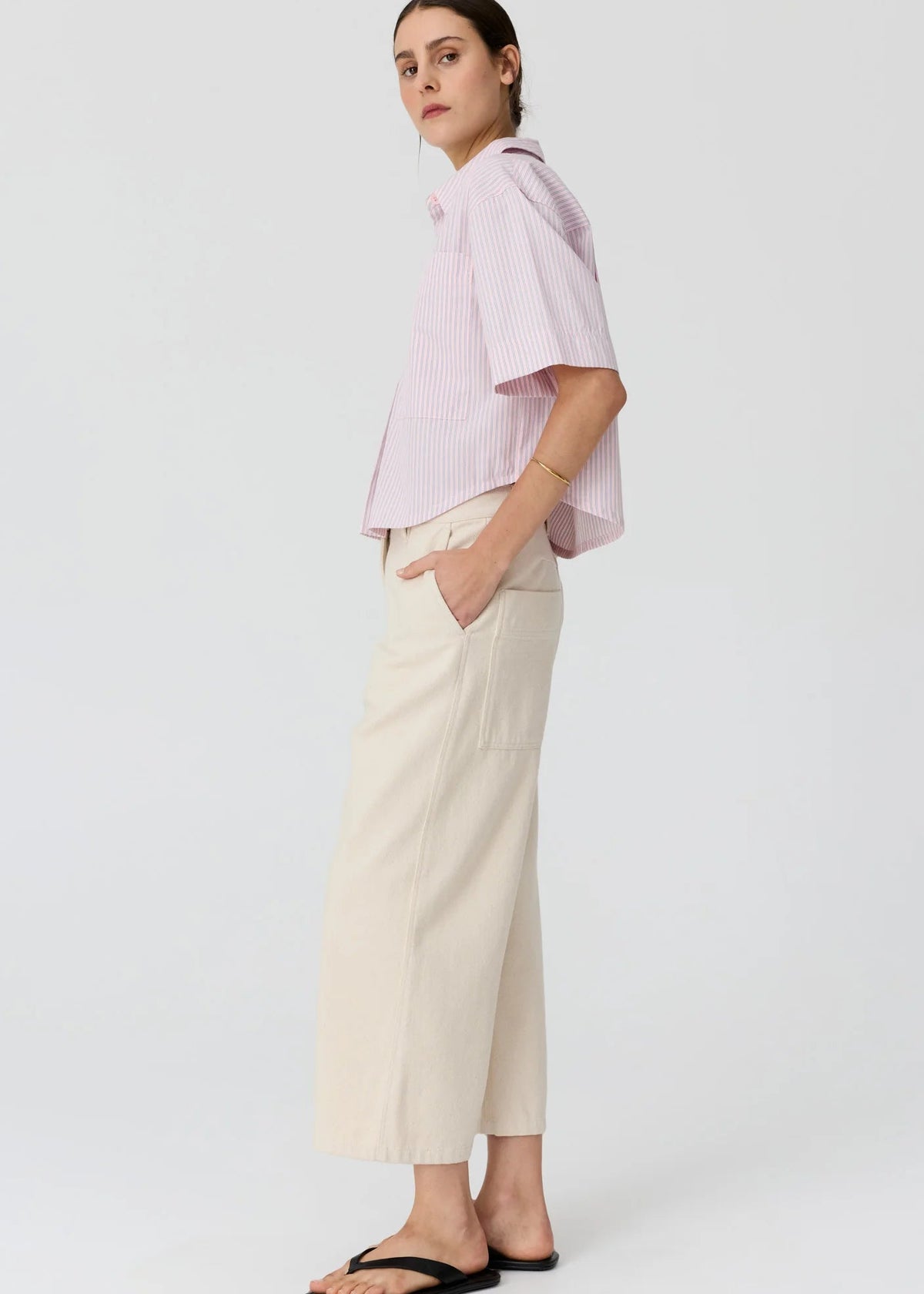 A woman modelling a light pink and white vertically striped short-sleeve button-up shirt paired with cream wide-leg cropped pants and black flip-flop sandals, set against a plain light grey studio background.