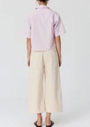 A woman modelling a light pink and white vertically striped short-sleeve button-up shirt paired with cream wide-leg cropped pants and black flip-flop sandals, set against a plain light grey studio background.