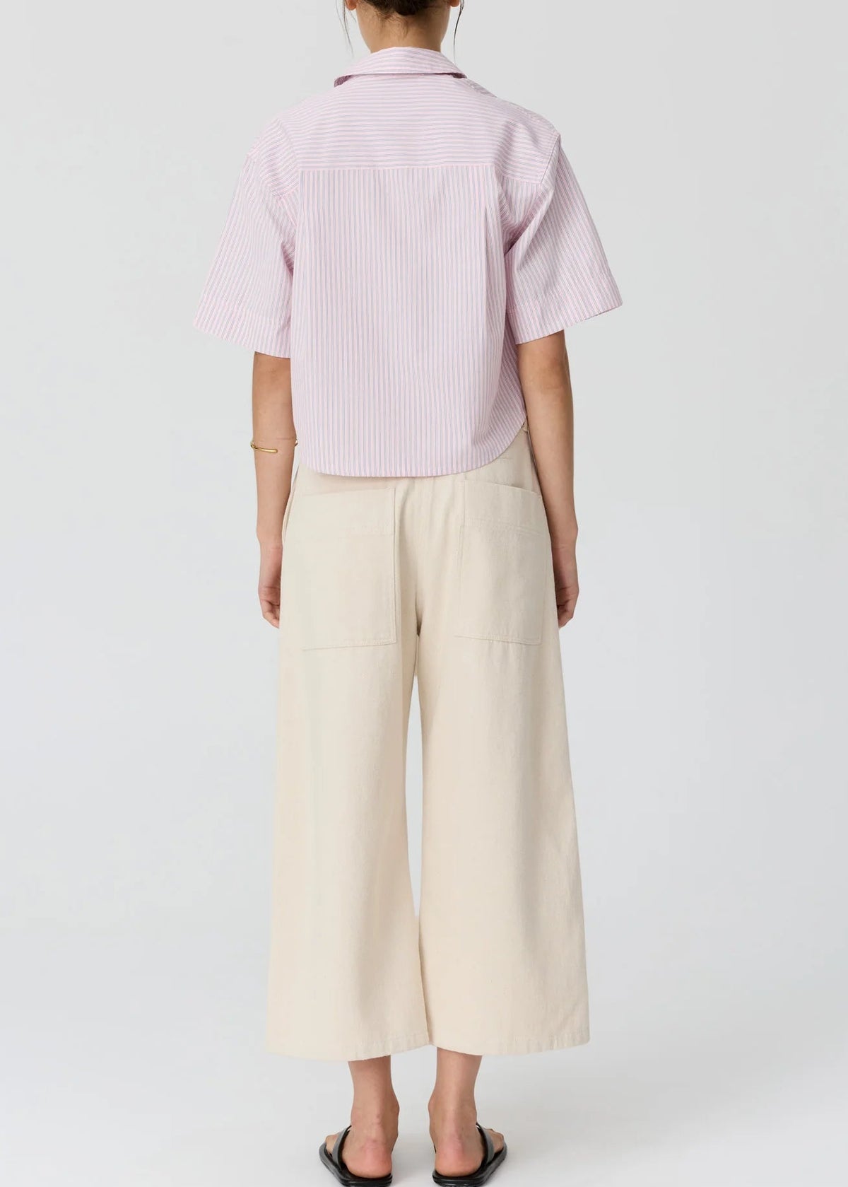 A woman modelling a light pink and white vertically striped short-sleeve button-up shirt paired with cream wide-leg cropped pants and black flip-flop sandals, set against a plain light grey studio background.
