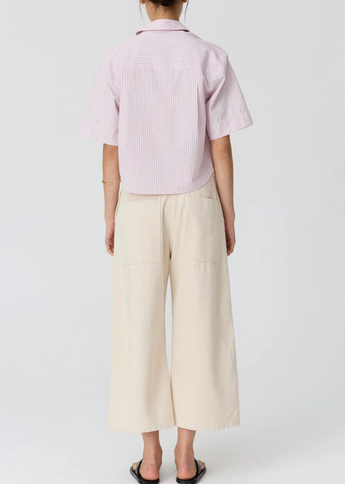 A woman modelling a light pink and white vertically striped short-sleeve button-up shirt paired with cream wide-leg cropped pants and black flip-flop sandals, set against a plain light grey studio background.