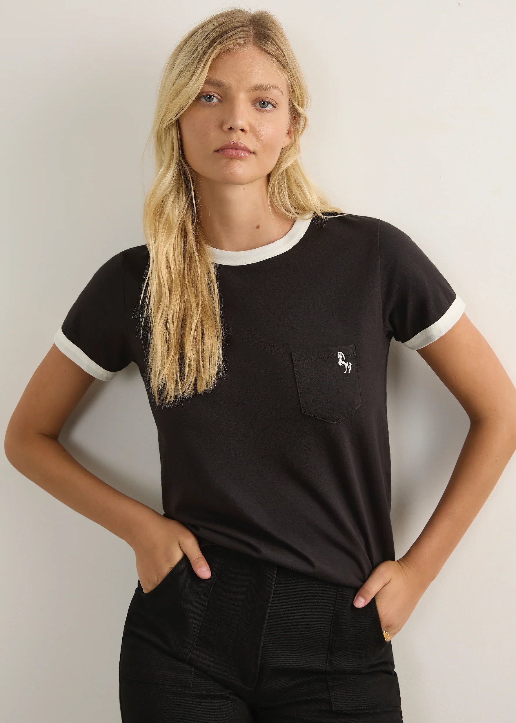 A blonde woman modelling a black ringer T-shirt with white trim on the crew neck and sleeve cuffs, featuring a left chest pocket with a small white embroidered logo.