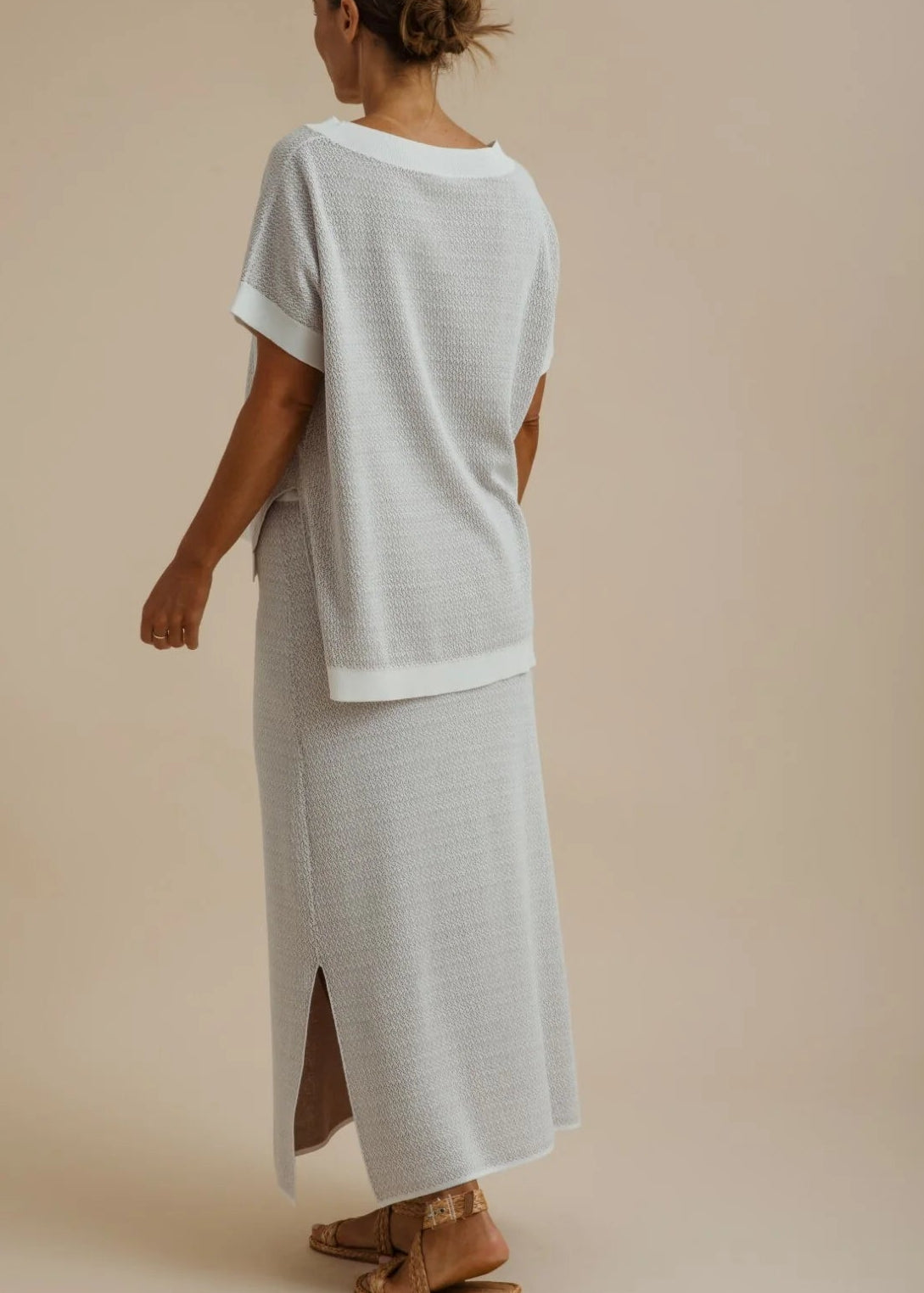 A woman shown from the back wearing a light grey short-sleeve top and matching long skirt with side slits, photographed against a plain warm beige studio background.