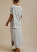 A woman shown from the back wearing a light grey short-sleeve top and matching long skirt with side slits, photographed against a plain warm beige studio background.