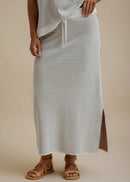 A person wearing a light gray textured maxi skirt with a side slit and drawstring waist, paired with brown leather sandals, against a warm beige studio background.