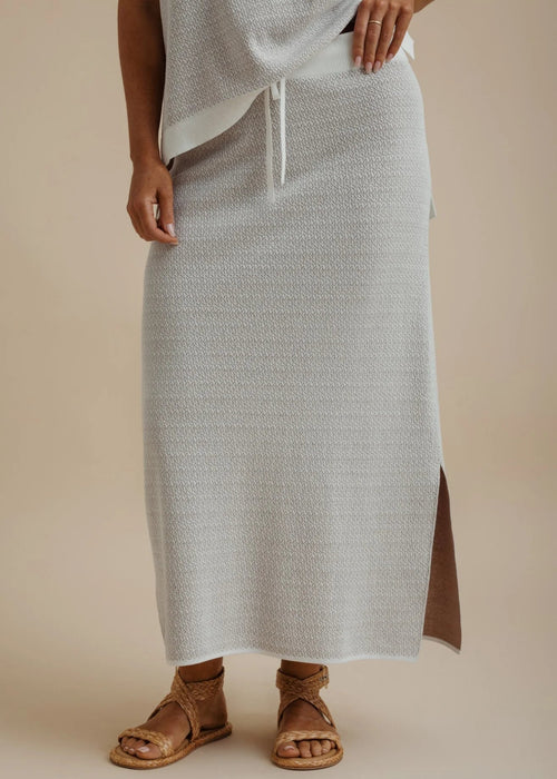 A person wearing a light gray textured maxi skirt with a side slit and drawstring waist, paired with brown leather sandals, against a warm beige studio background.