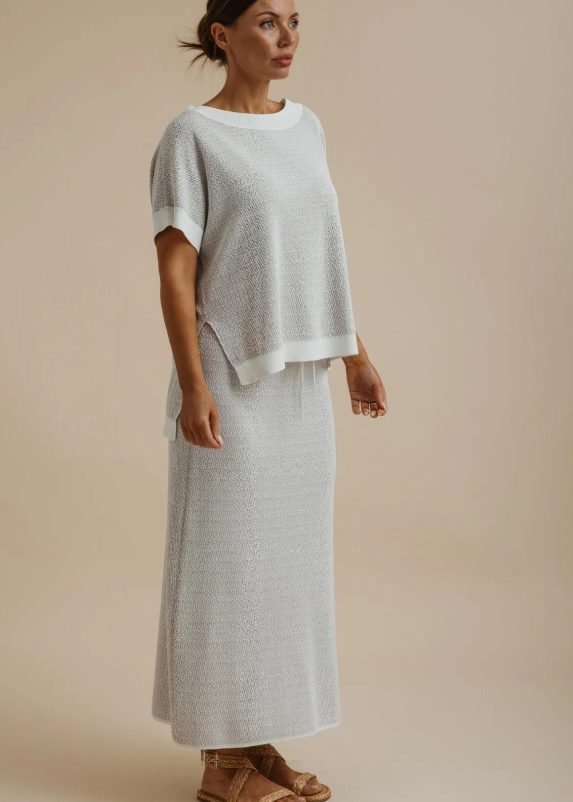 A woman shown from the back wearing a light grey short-sleeve top and matching long skirt with side slits, photographed against a plain warm beige studio background.