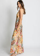 A woman modelling a long spaghetti-strap maxi dress with a multicolour collage print of vintage postcards, flowers, and illustrations, set against a plain light grey studio background.