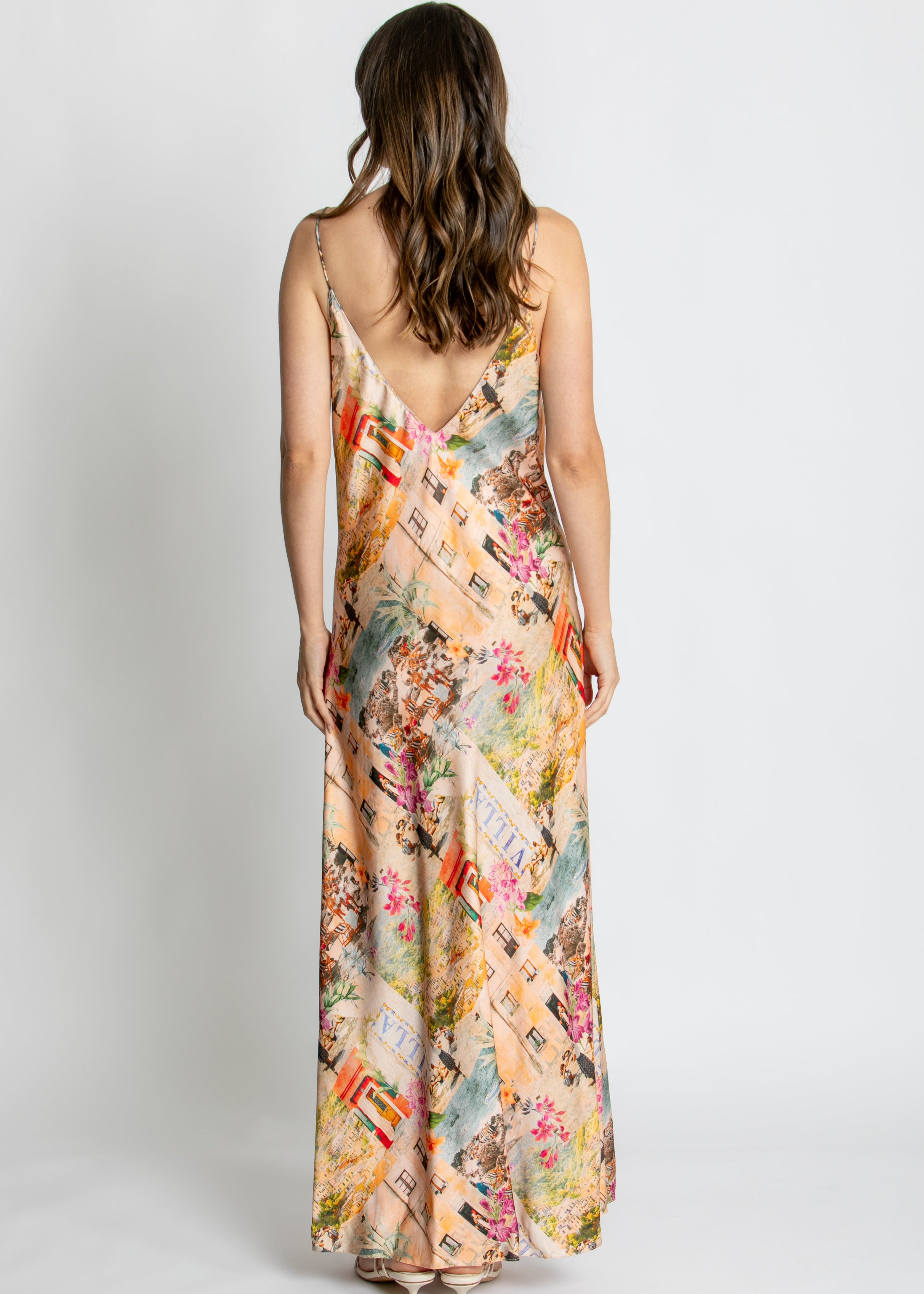 A woman modelling a long spaghetti-strap maxi dress with a multicolour collage print of vintage postcards, flowers, and illustrations, set against a plain light grey studio background.