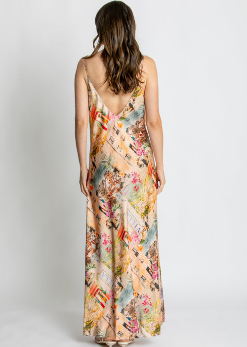 A woman modelling a long spaghetti-strap maxi dress with a multicolour collage print of vintage postcards, flowers, and illustrations, set against a plain light grey studio background.