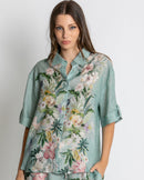 A woman modelling a matching two-piece lounge set consisting of a short-sleeve button-up shirt and shorts in a sage green base with a vibrant pink, white, and yellow floral print, set against a plain light grey background.