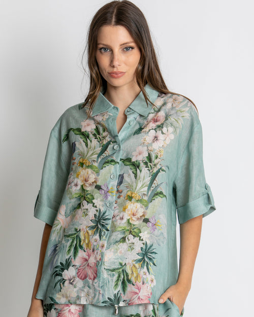 A woman modelling a matching two-piece lounge set consisting of a short-sleeve button-up shirt and shorts in a sage green base with a vibrant pink, white, and yellow floral print, set against a plain light grey background.