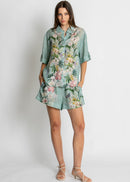 A woman modelling a matching two-piece lounge set consisting of a short-sleeve button-up shirt and shorts in a sage green base with a vibrant pink, white, and yellow floral print, set against a plain light grey background.