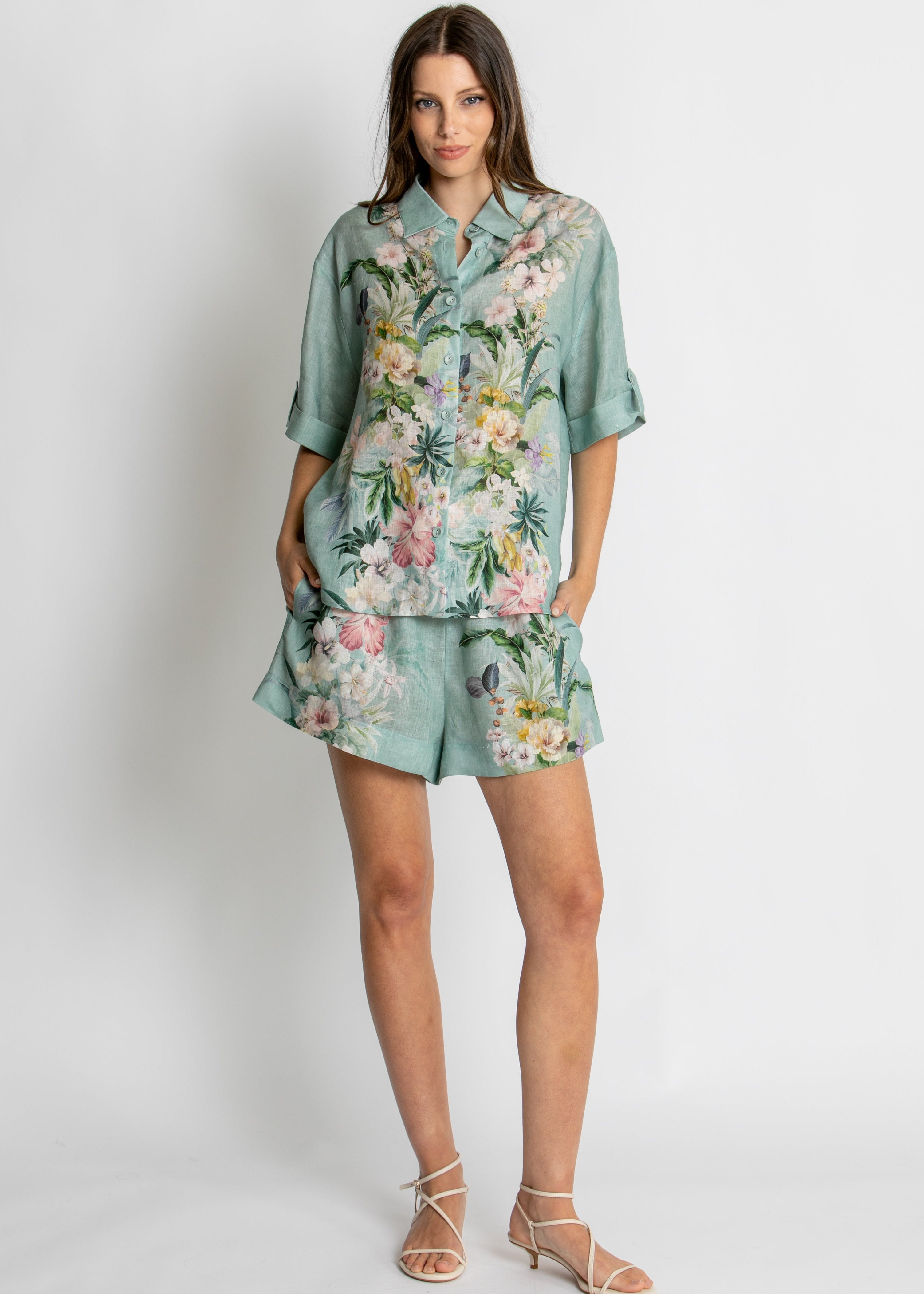 A woman modelling a matching two-piece lounge set consisting of a short-sleeve button-up shirt and shorts in a sage green base with a vibrant pink, white, and yellow floral print, set against a plain light grey background.