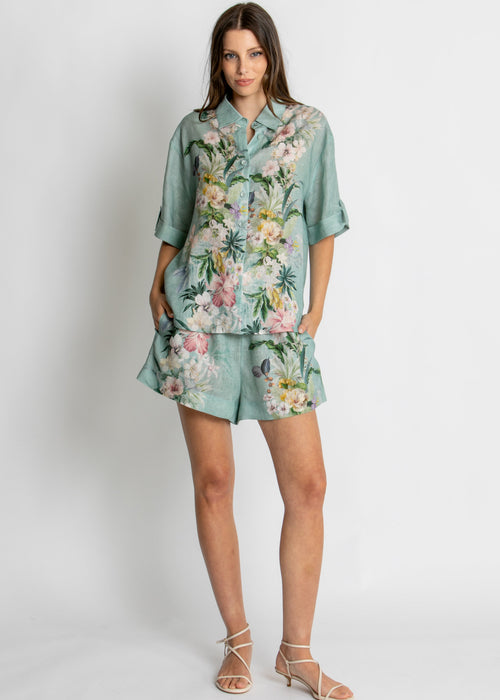 A woman modelling a matching two-piece lounge set consisting of a short-sleeve button-up shirt and shorts in a sage green base with a vibrant pink, white, and yellow floral print, set against a plain light grey background.
