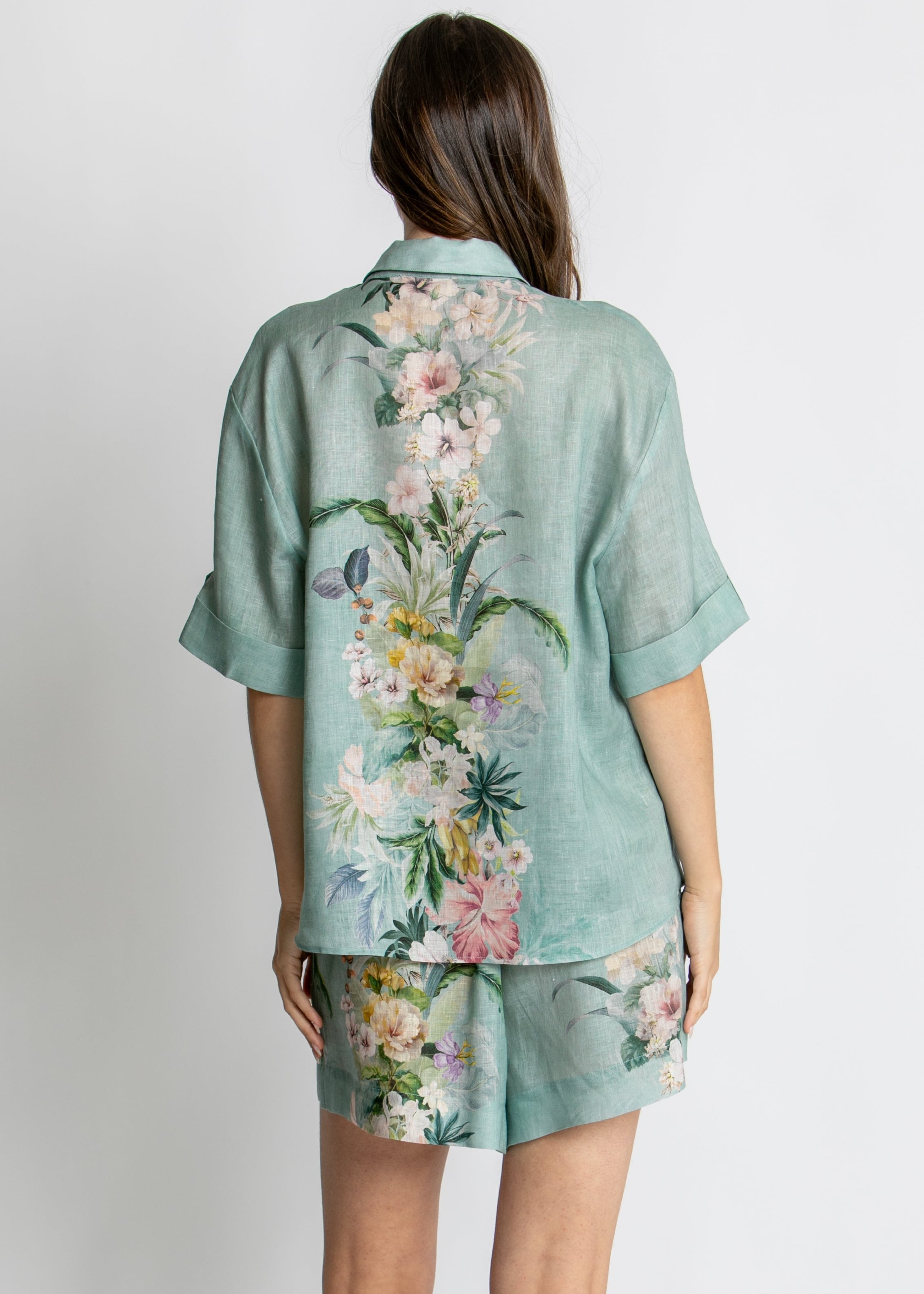 A woman modelling a matching two-piece lounge set consisting of a short-sleeve button-up shirt and shorts in a sage green base with a vibrant pink, white, and yellow floral print, set against a plain light grey background.