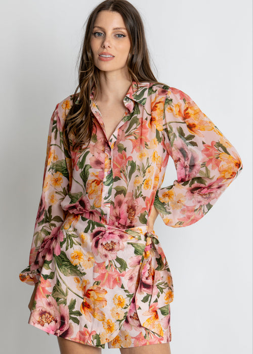 A woman modelling a long-sleeve floral shirt dress with a matching tie belt, featuring a vibrant pink, orange, and yellow floral pattern on a light background, set against a plain light grey studio backdrop.