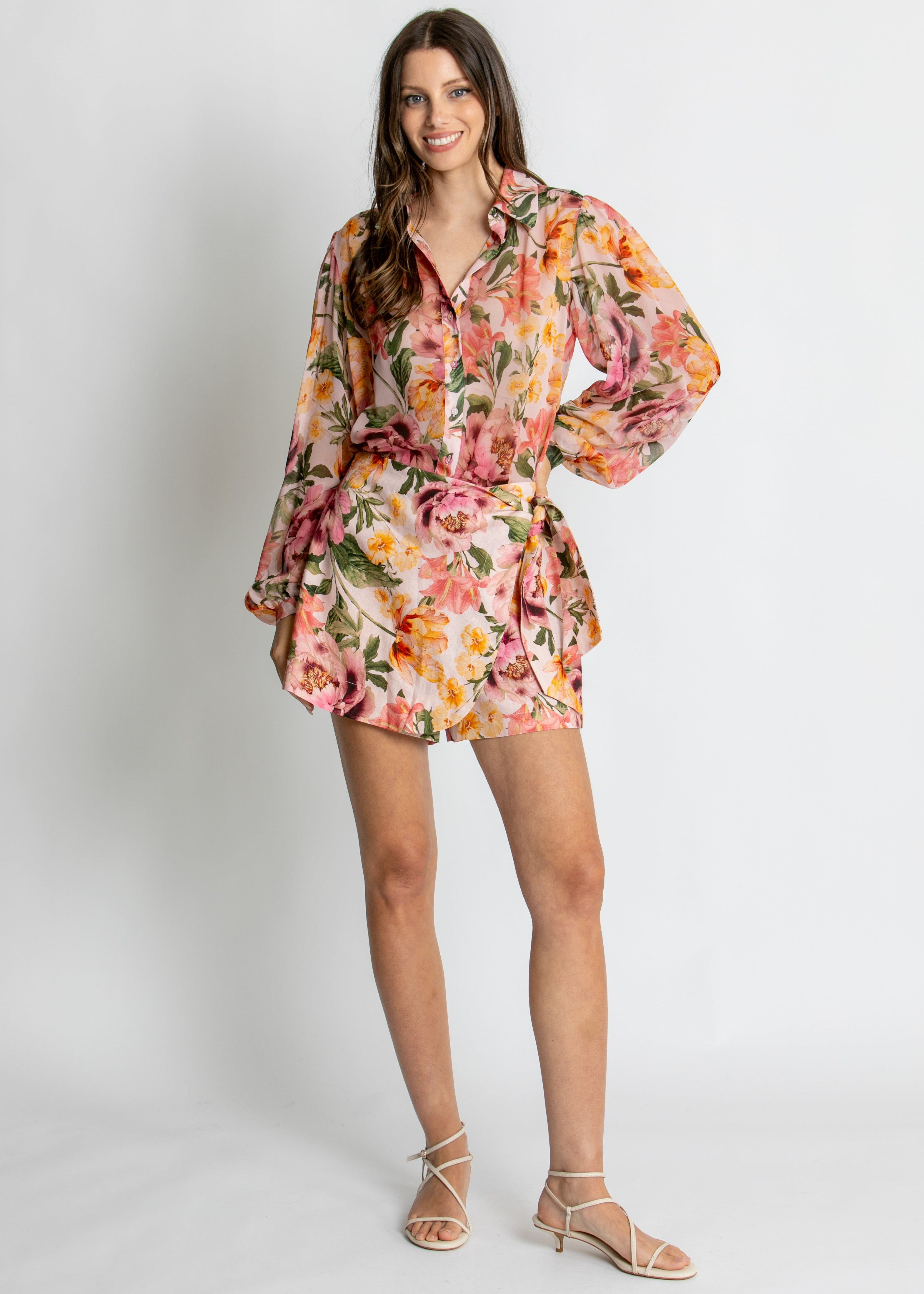 A woman modelling a long-sleeve floral shirt dress with a matching tie belt, featuring a vibrant pink, orange, and yellow floral pattern on a light background, set against a plain light grey studio backdrop.