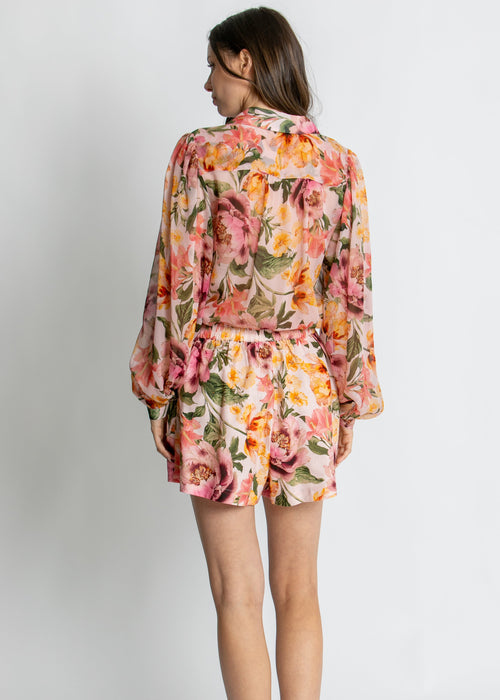 A woman modelling a long-sleeve floral shirt dress with a matching tie belt, featuring a vibrant pink, orange, and yellow floral pattern on a light background, set against a plain light grey studio backdrop.