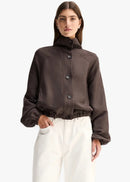 A model wearing a dark brown cropped button-up jacket with a high mock neck and gathered cuffs, paired with white trousers, against a light grey background.